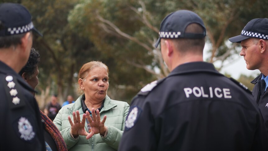 A woman in a light green jacket talks to police officers in dark jackets outside with trees in background. 