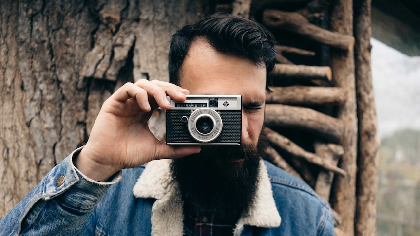 A man with coiffed black hair and a fashionable beard holds a vintage camera.