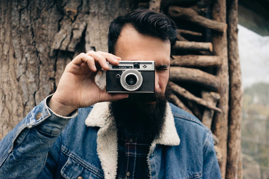 A man with coiffed black hair and a fashionable beard holds a vintage camera.