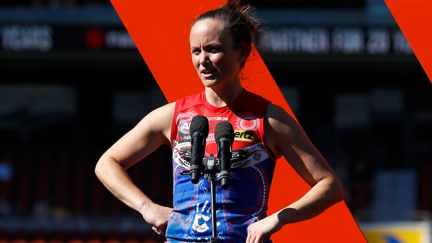 AFLW star Daisy Pearce stands on stage near a microphone with hands on hips after her team loses a grand final.