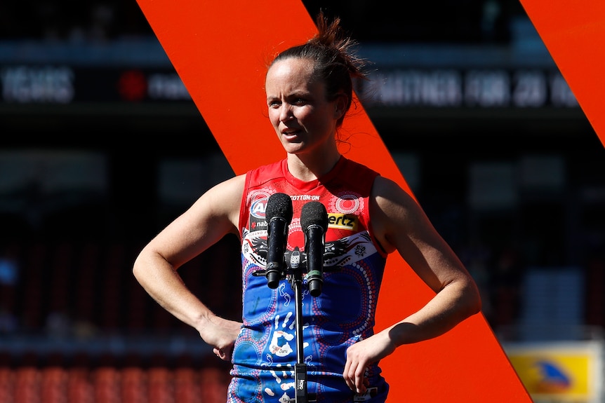 AFLW star Daisy Pearce stands on stage near a microphone with hands on hips after her team loses a grand final.