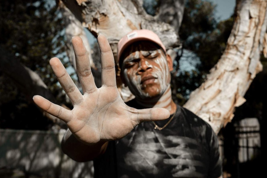 Indigenous man standing in front of a paperbark tree, his face is painted with white clay lines