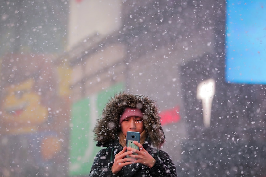 A woman in a big furry hooded jacket texts on her phone while standing in snow 