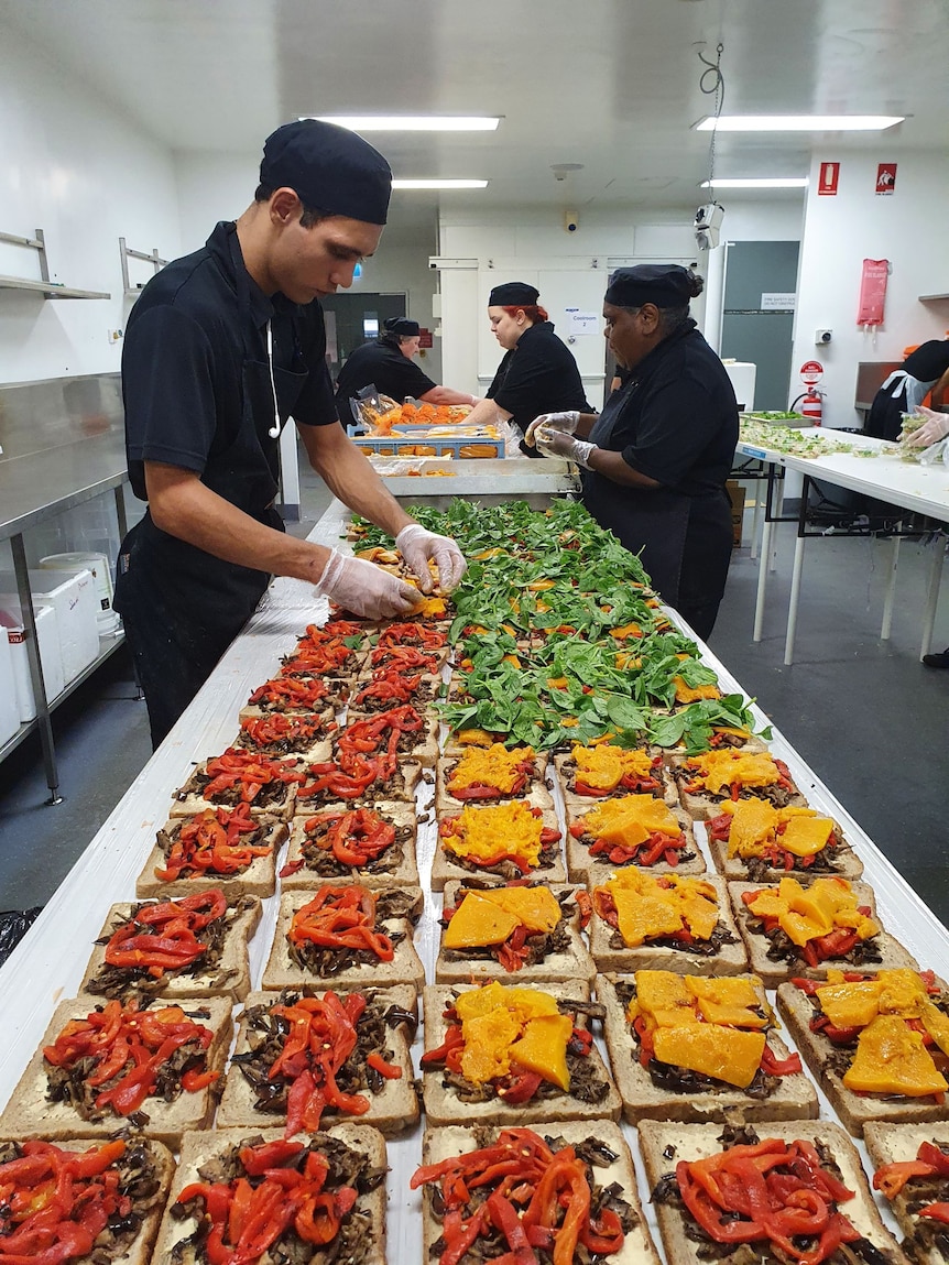 Chefs stand at a long table in a kitchen, adding finishing touches to a line of meals.