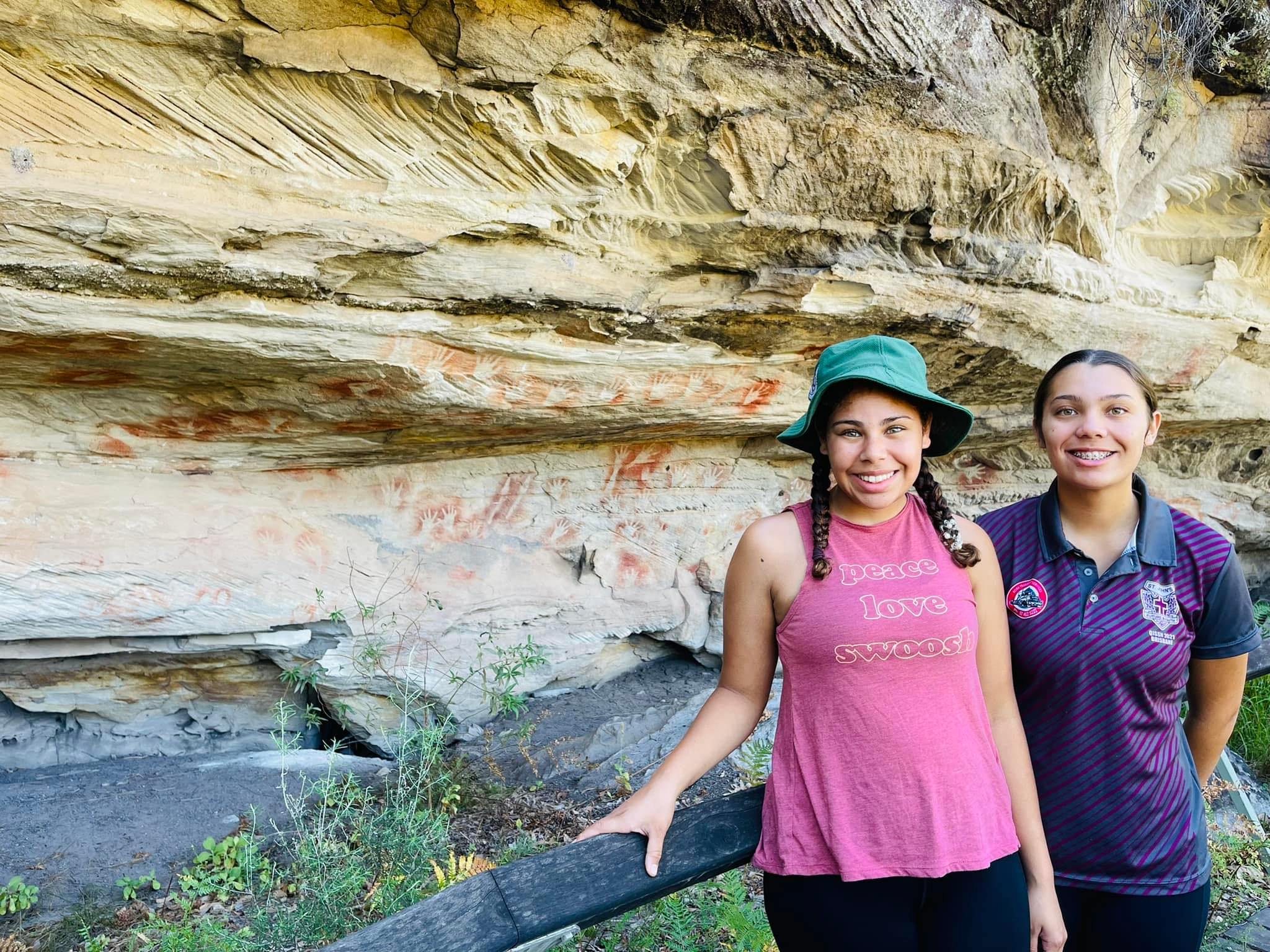 Two girls smiling at the camera in front of Aboriginal rock art. 