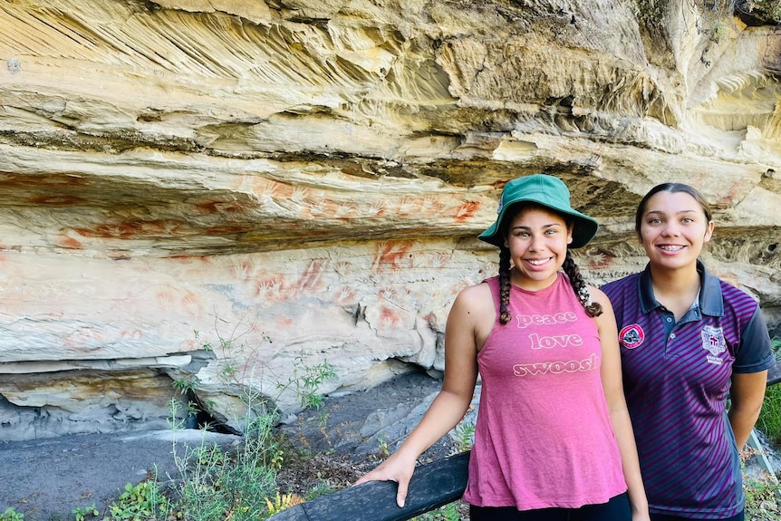 Two girls smiling at the camera in front of Aboriginal rock art. 