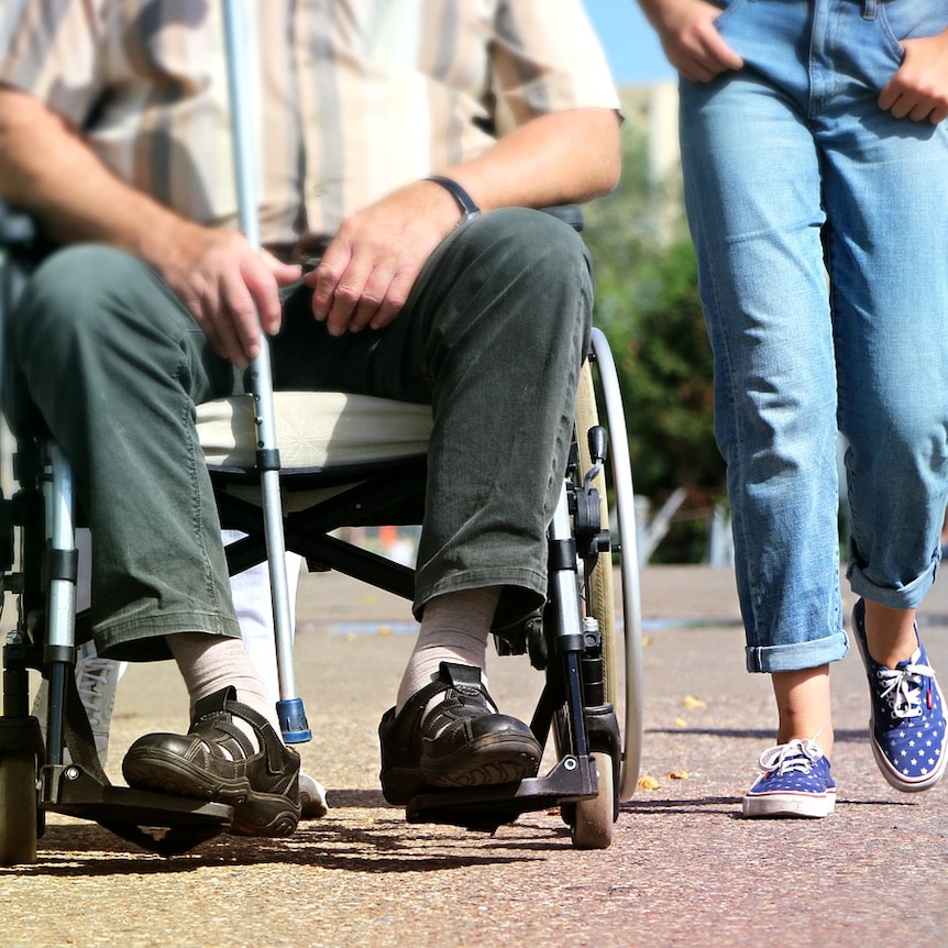 A man in a wheelchair holding a cane next to someone walking and wearing jeans.