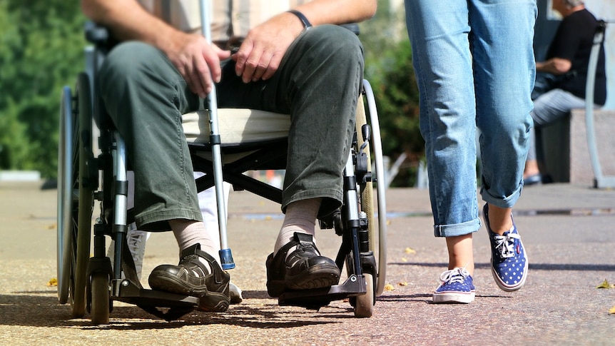 A man in a wheelchair holding a cane next to someone walking and wearing jeans.