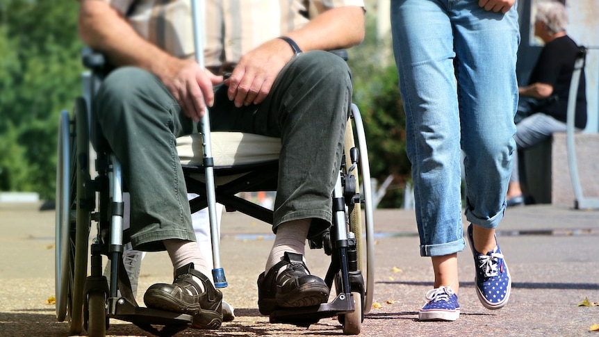 A man in a wheelchair holding a cane next to someone walking and wearing jeans.