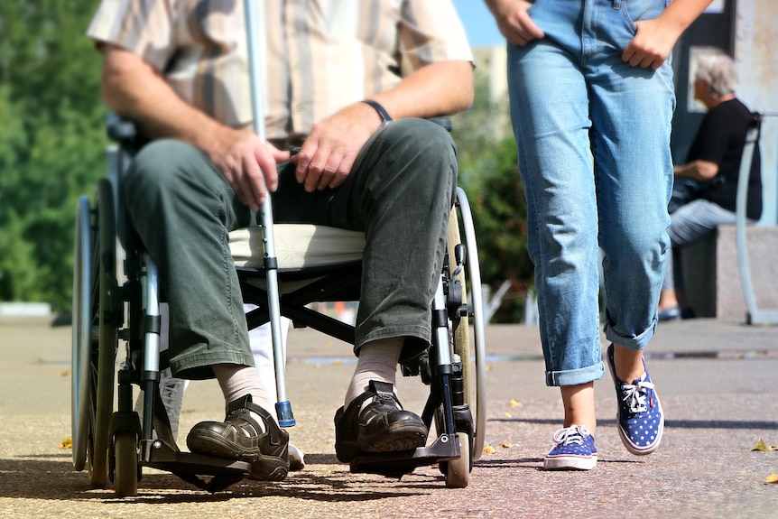 A man in a wheelchair holding a cane next to someone walking and wearing jeans.