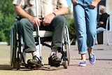 A man in a wheelchair holding a cane next to someone walking and wearing jeans.