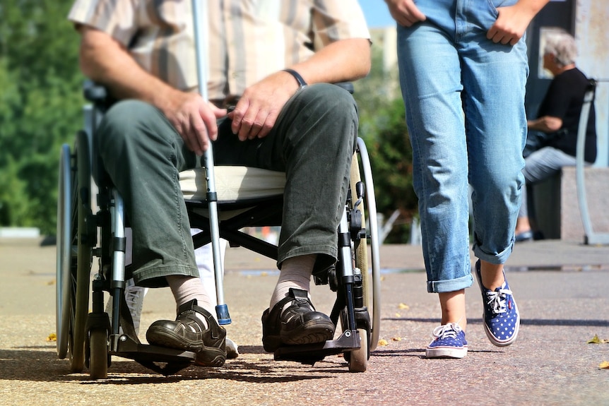 A man in a wheelchair holding a cane next to someone walking and wearing jeans.