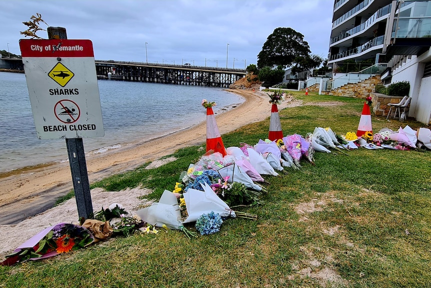A row of flower bouquets near a river bank, with a sign saying 'sharks beach closed'