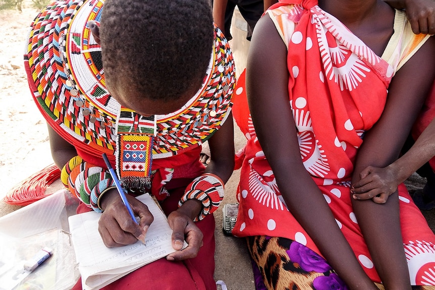 A girl wearing colourful beadwork around her neck writes in a notebook with a pencil.
