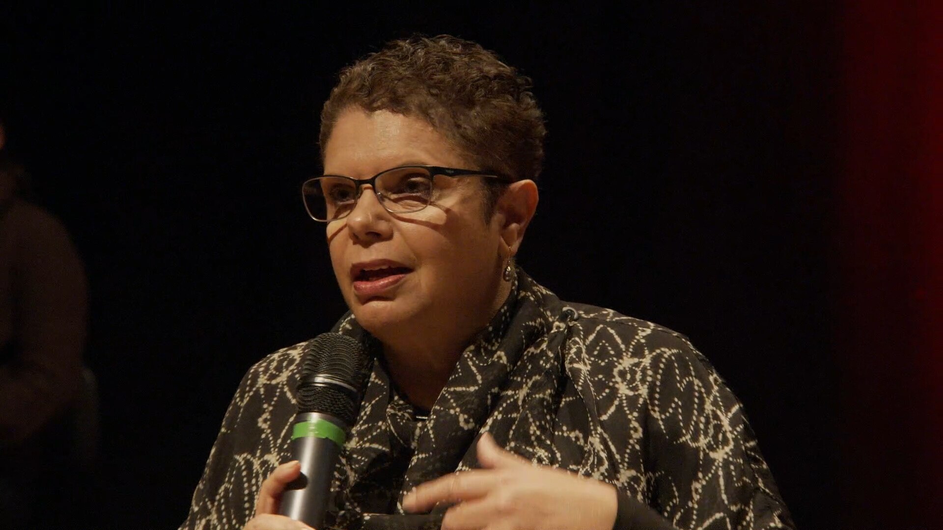 Soprano and composer Deborah Cheetham talks into a microphone in a darkened theatre.