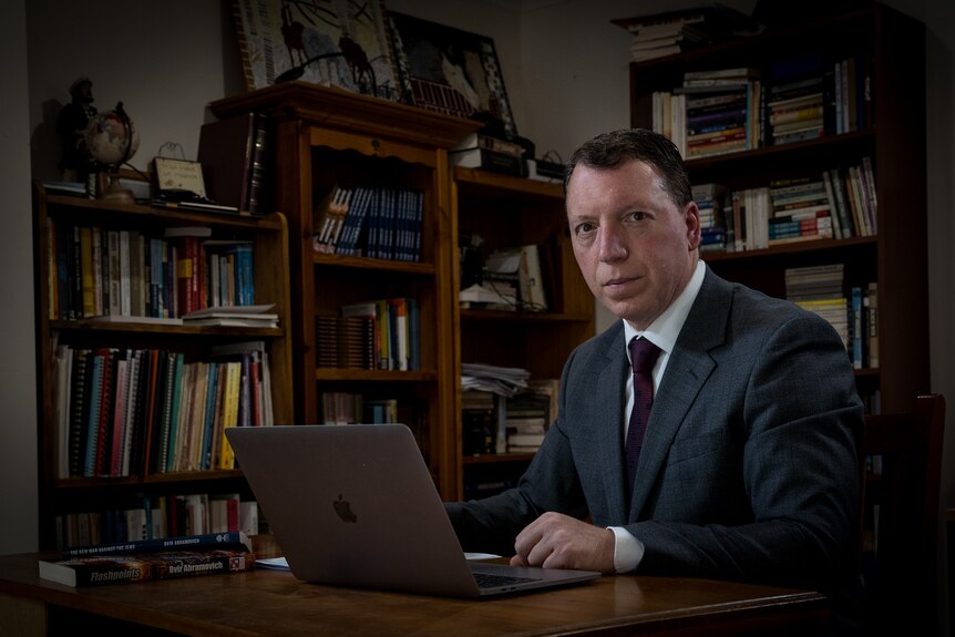 A dark-haired man in a dark suit sits in a library, operating a laptop.