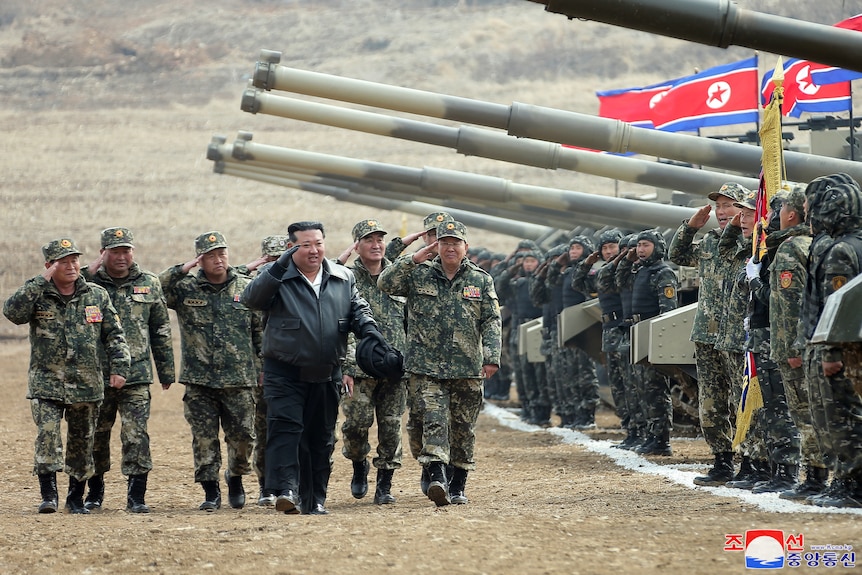 Kim Jong Un salutes as he walks with men dressed in military uniform past tanks and other men in uniform.