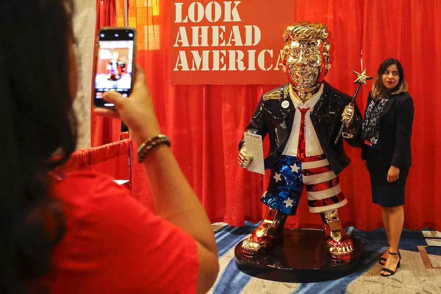 An African-American woman poses next to golden statue at a conference as another woman takes photo.