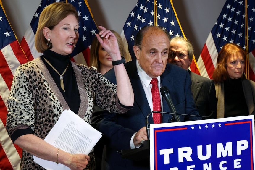 A tall woman in a cheetah-print suit and a tired-looking man in a blue suit stand behind a Trump-Pence lectern.