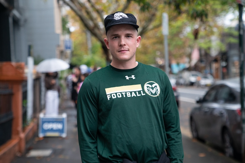 A man wearing a dark green football t-shirt and a black cap with a white logo stands on a busy sidewalk.