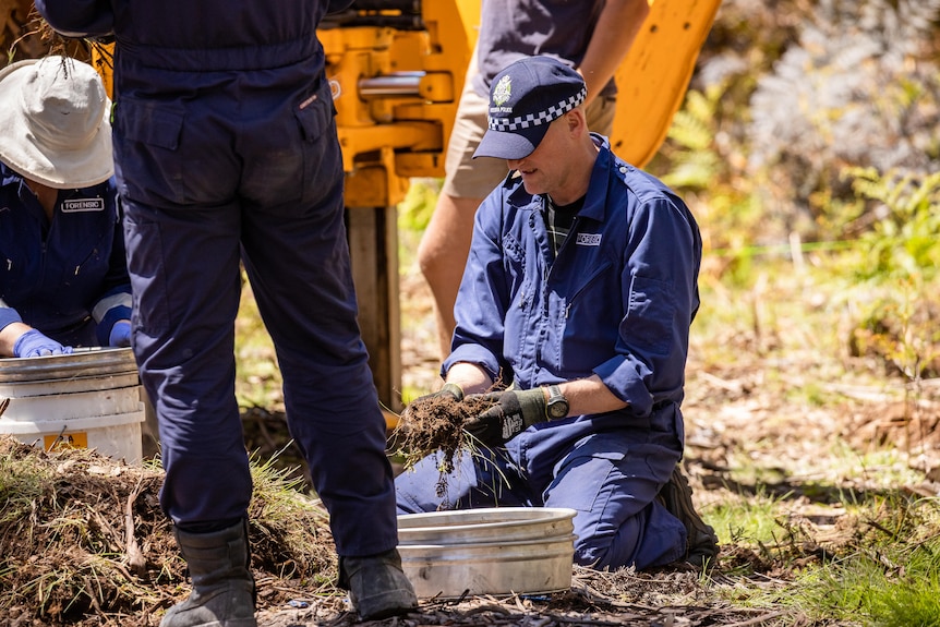 Man in police officers uniform kneels on ground clutching clump of dirt, while in foreground legs wearing blue pants seen.