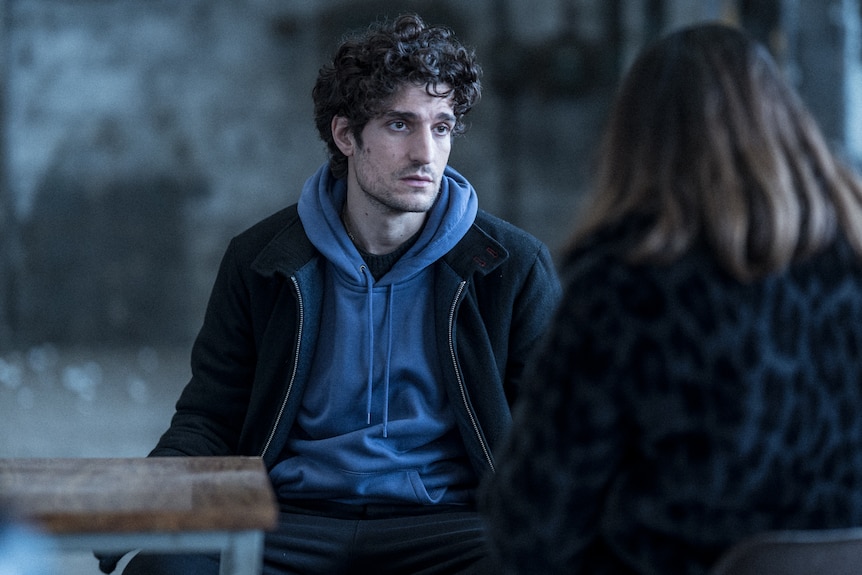 Louis Garrel, a 30-something white man in a blue hoodie and dark jacket sits in a warehouse looking morose.