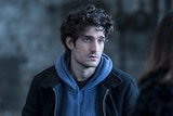 Louis Garrel, a 30-something white man in a blue hoodie and dark jacket sits in a warehouse looking morose.