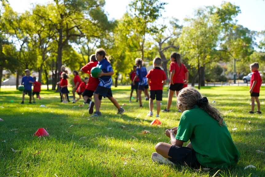 A young girl in green shirt sits on the grass in the foreground while other kids are playing dodge ball in the background. 