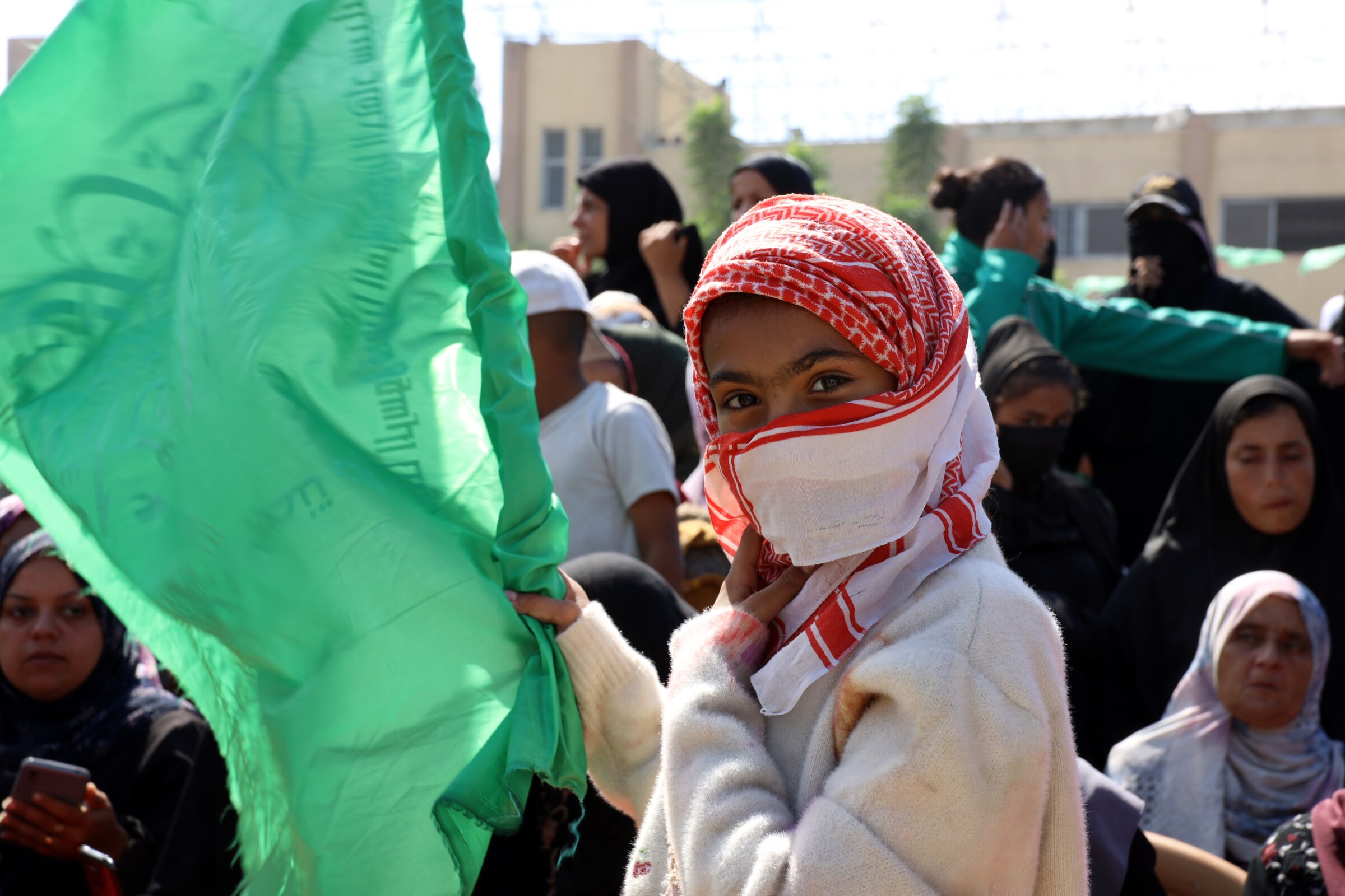 A Palestinian girl waves a flag as a crowd gathers around her waving their arms.