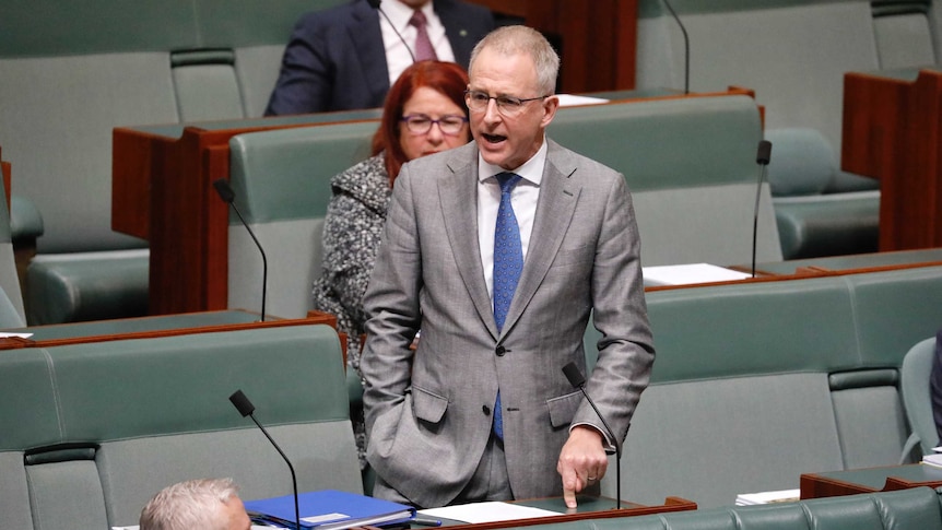 A white man with grey hair and glasses in a grey suit stands up in the house of representatives speaking