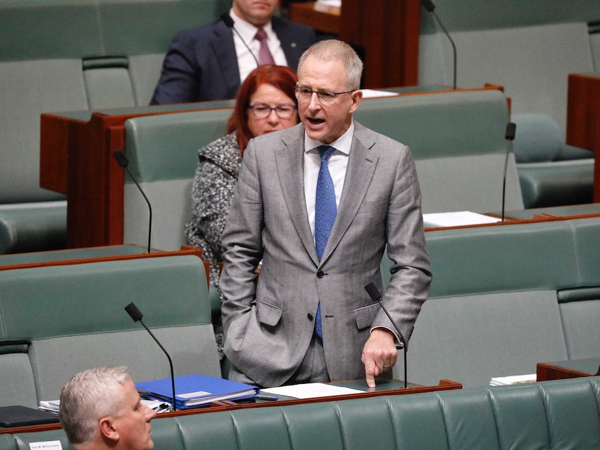 A white man with grey hair and glasses in a grey suit stands up in the house of representatives speaking