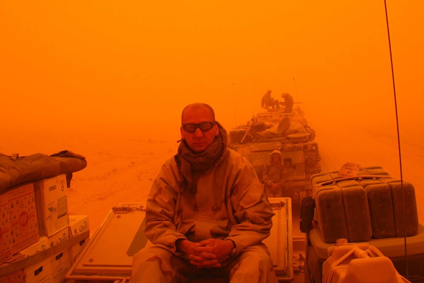 Orange-hued photograph of a man in military gear and goggles on the back of military tank.