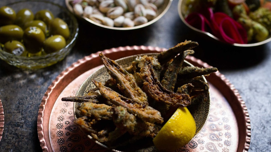 Fried okra with a wedge of lemon in a small bowl on slate bench, alongside other mezze.