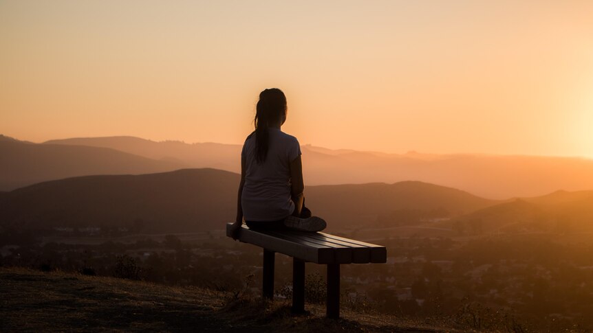 A woman sits on a bench and watches the sun set over hills in the distance.