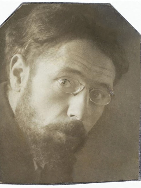 A sepia photograph of a man with thin-rimmed glasses and a short beard, gazing at the camera.