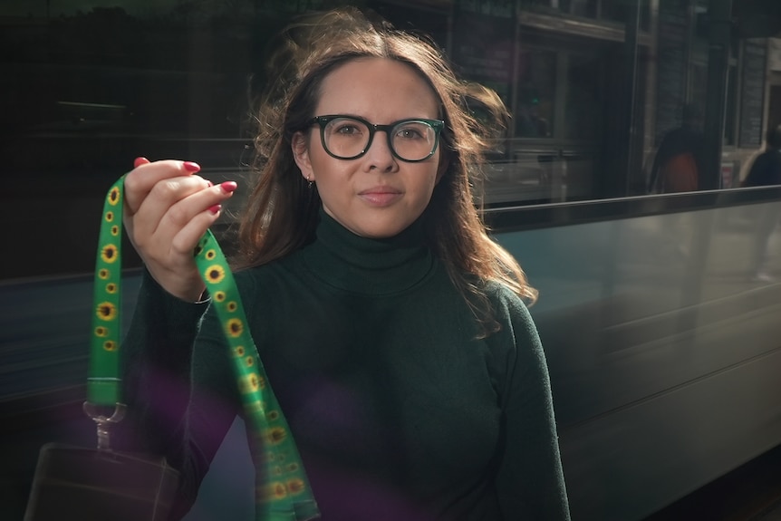 A young white woman with long brown hair standing int he street. She's holding a bright green lanyard with sunflowers on it
