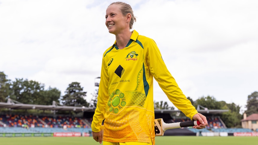 Meg Lanning smiles and holds a cricket bat while wearing Australia's gold ODI strip