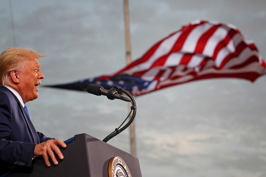 Donald Trump speaking on a stage with a US flag flapping behind him