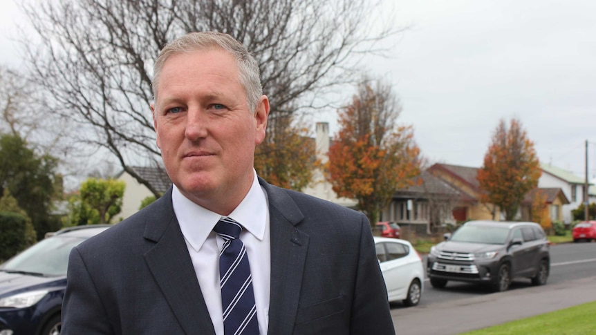 A man in a suit stands on a street and looks at the camera.