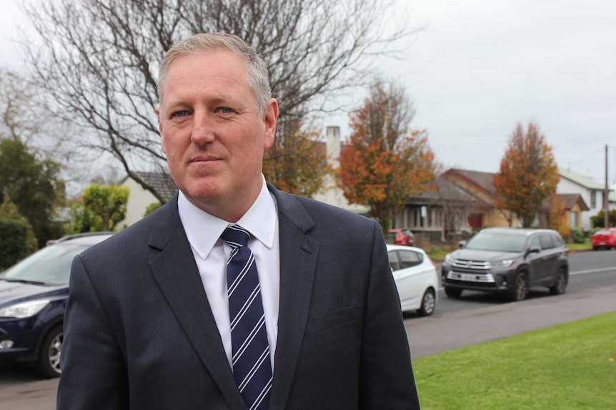 A man in a suit stands on a street and looks at the camera.