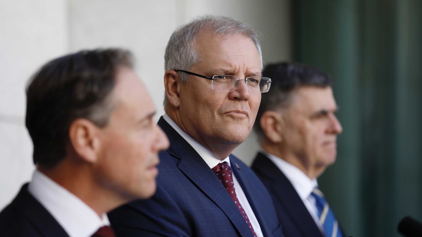 Scott Morrison looks into the distance, standing beside Greg Hunt and Brendan Murphy at podia with microphones