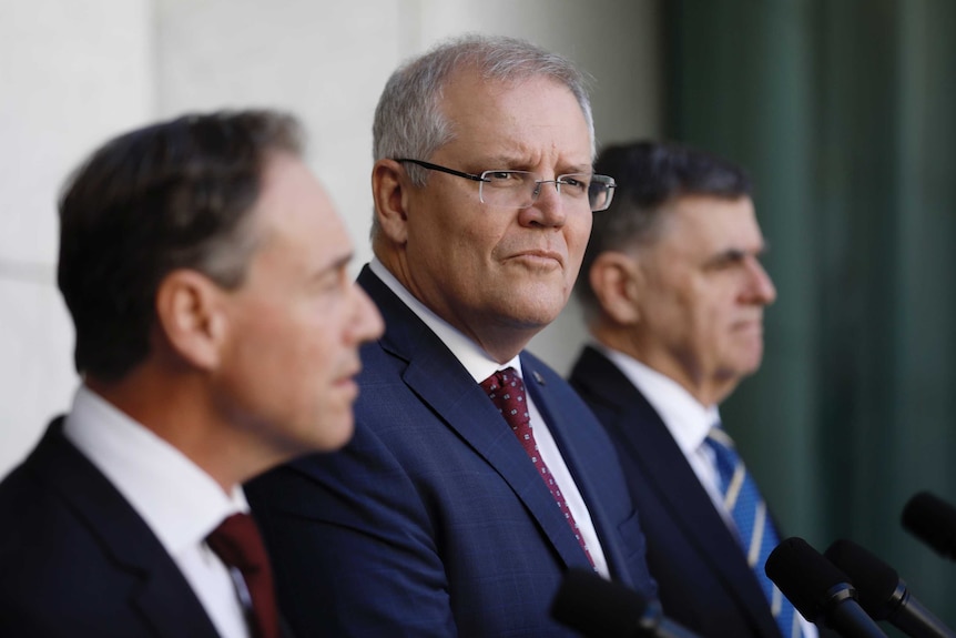 Scott Morrison looks into the distance, standing beside Greg Hunt and Brendan Murphy at podia with microphones