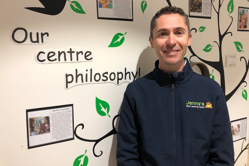 Smiling man, short black hair, wears black branded hoodie, stands in front of a wall with tree, birds, our centre philiosophy.