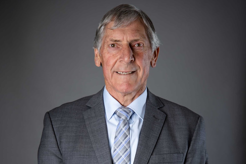 An older man in a grey suit with a  blue tie smiles at the camera