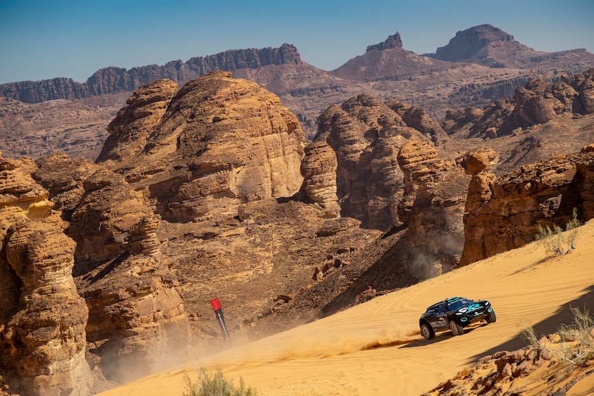 A car drives up a hill of sand with a scenic hilly, rocky backdrop behind it