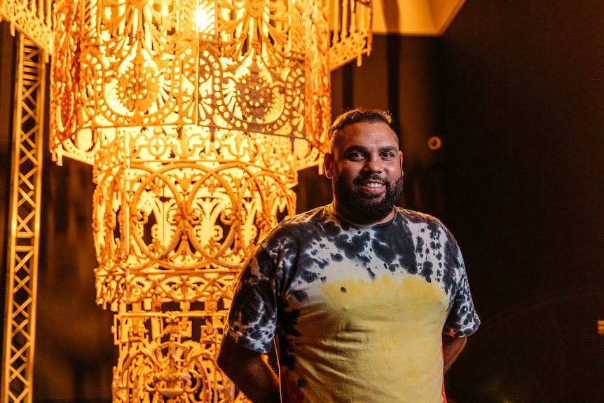 Warrang man with short dark hair and beard smiles wearing a tie-dyed Aboriginal flag print shirt beside large yellow chandelier