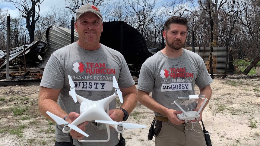 Two men hold drones in front of  burnt out shed