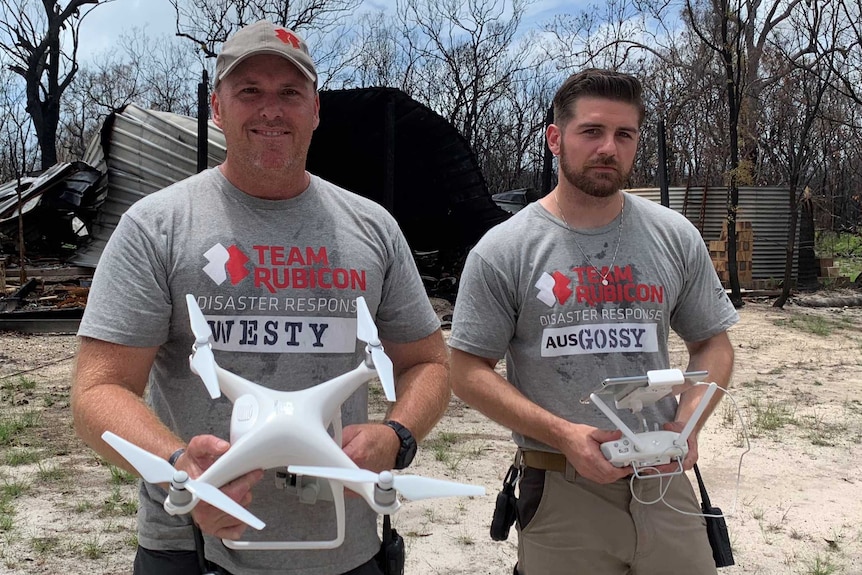 Two men hold drones in front of  burnt out shed