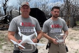 Two men hold drones in front of  burnt out shed