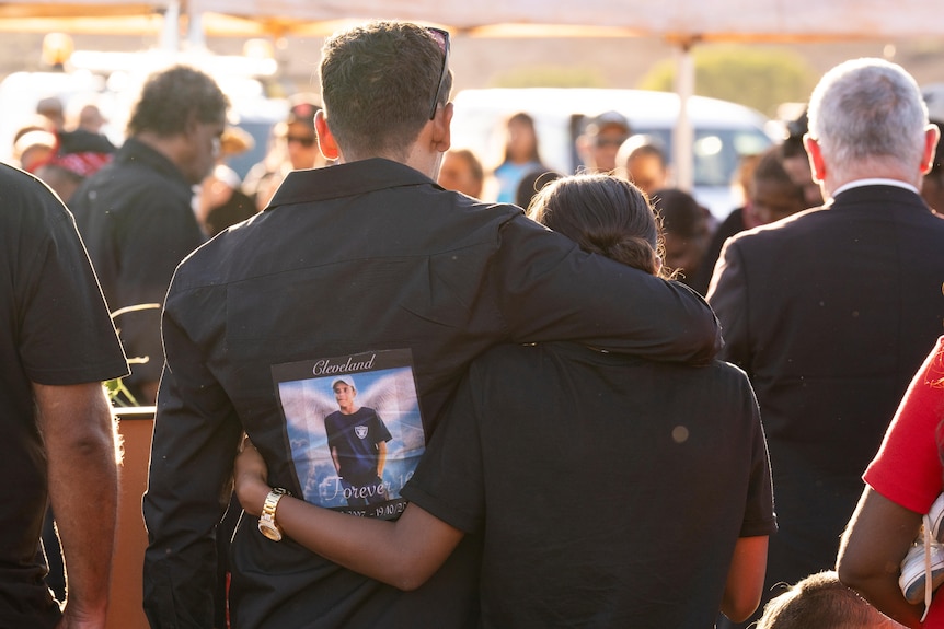 Two people dressed in black embrace at a funeral. 